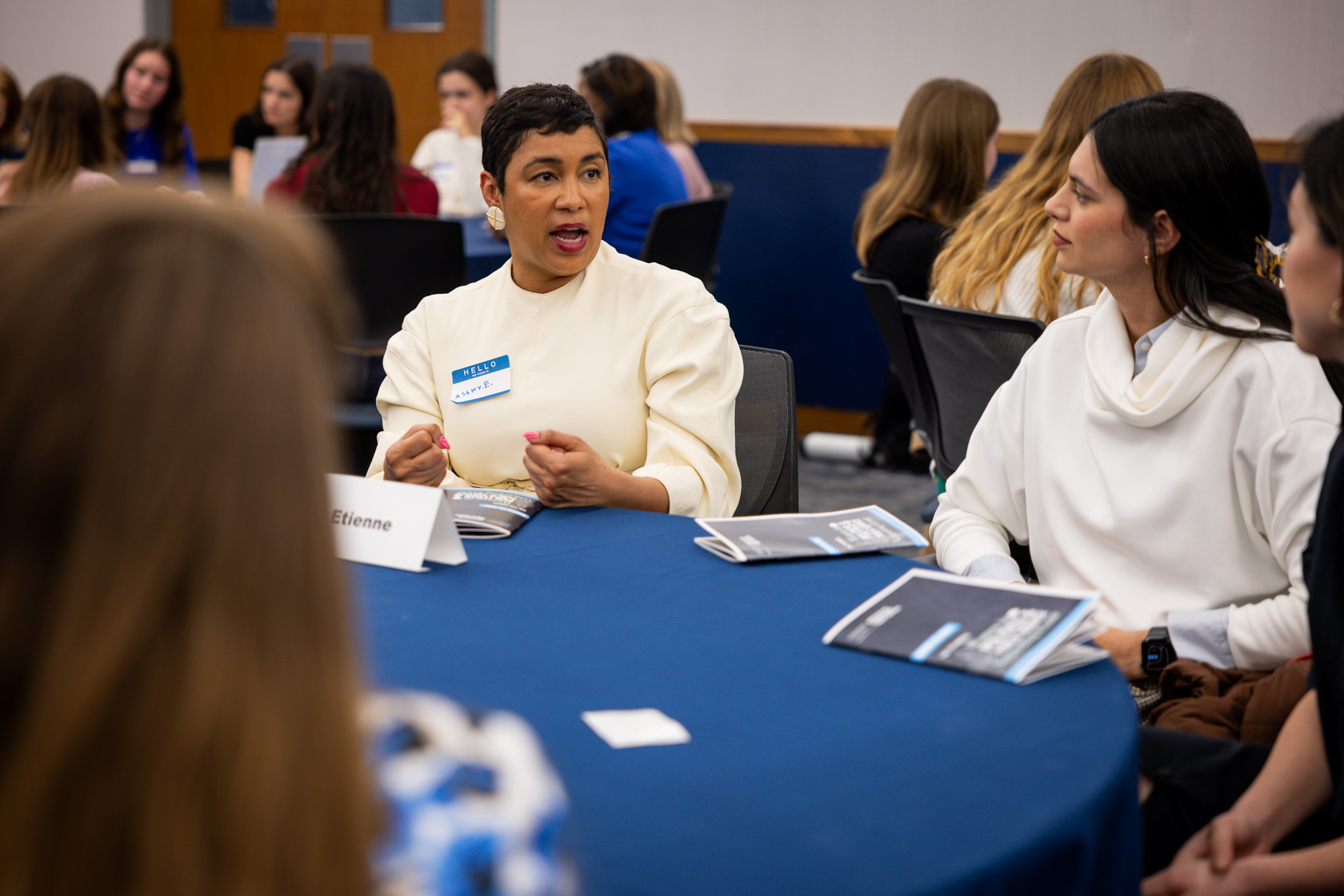 Ashely Etienne speaks to student at table. Tables of students in background.